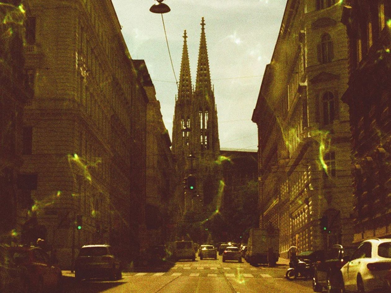 City street with tall buildings and a prominent spire under an overcast sky, with subtle gemstone green effects across the frame.